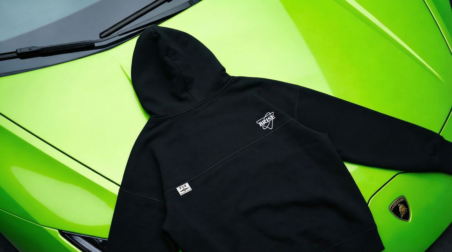 Brise hoodie in triple black, back view, laid on the green hood of a Lamborghini: white shield logo upper right, ブリズ patch on the yoke